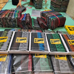 Puede incluir: Un surtido de telas plegadas, algunas en cajas, con diversos patrones y colores. Los tejidos incluyen rayas y diseños geométricos en tonos verdes, azules, rojos y negros. Algunos artículos están empaquetados en plástico transparente.