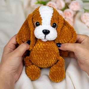 May include: A plush, crocheted dog toy in shades of brown and white. The dog has floppy ears, a white stripe down its head, and a white muzzle. The toy is held by hands, and the background includes soft fabric and flowers.