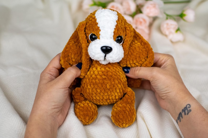 May include: A plush, crocheted dog toy in shades of brown and white. The dog has floppy ears, a white stripe down its head, and a white muzzle. The toy is held by hands, and the background includes soft fabric and flowers.