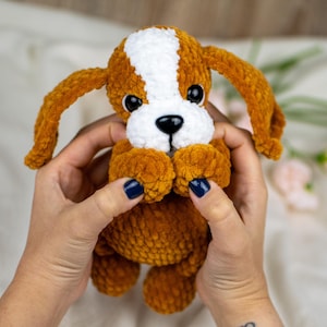 May include: A handmade plush puppy with brown and white fur, large black eyes, and long floppy ears. The puppy is held in a person's hands. The plush toy is a soft, cuddly item.