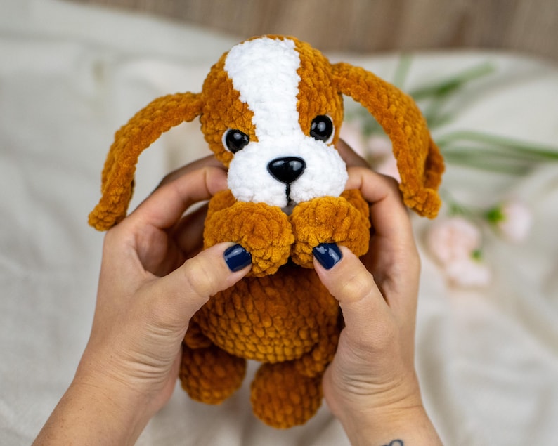 May include: A handmade plush puppy with brown and white fur, large black eyes, and long floppy ears. The puppy is held in a person's hands. The plush toy is a soft, cuddly item.