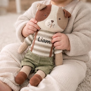 Puede incluir: Un peluche de perro tejido con detalles en marrón y crema. El perro lleva una camisa a rayas, pantalones verdes y tirantes marrones. El nombre "Liam" está impreso en la camisa. El juguete está sostenido por una persona que lleva un suéter color crema.