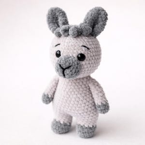 Puede incluir: Una llama de ganchillo hecha a mano en gris suave y blanco. El animal de peluche tiene ojos grandes y oscuros, orejas grises y un hocico gris. La llama está de pie sobre una superficie blanca.