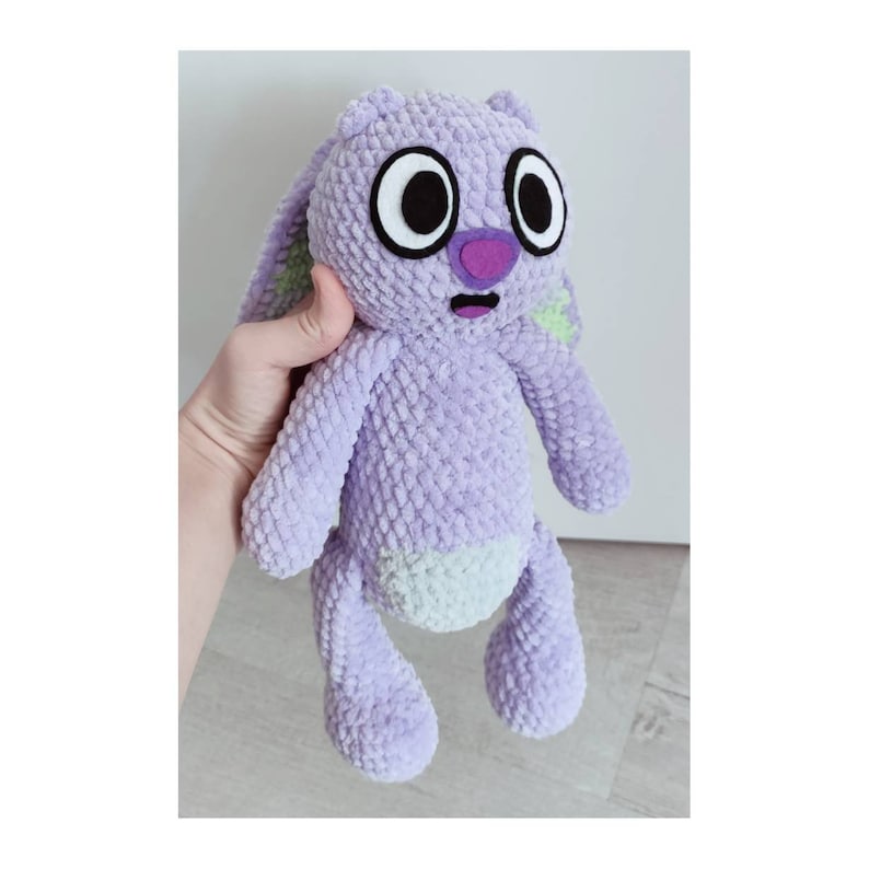 May include: A plush toy made of light purple textured yarn featuring long ears with lime green accents, a dark purple nose, and large white eyes with black pupils. The stuffed animal has a light-colored belly patch and a small open mouth expression.