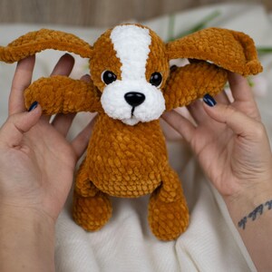 May include: A handmade plush dog toy in shades of brown and white. The dog has large, floppy ears, a white stripe down its head, and black eyes. The toy is made of a soft, textured material.