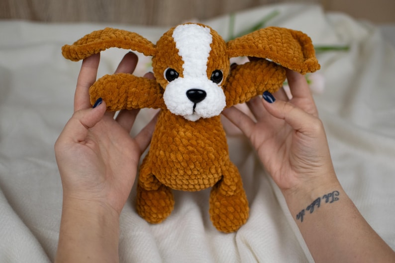 May include: A handmade plush dog toy in shades of brown and white. The dog has large, floppy ears, a white stripe down its head, and black eyes. The toy is made of a soft, textured material.
