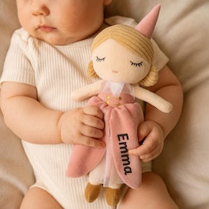 Puede incluir: Una muñeca de peluche suave con temática de princesa. La muñeca tiene piel clara, ojos cerrados y cabello rubio. Viste un vestido rosa con detalles dorados y un sombrero en forma de cono a juego. El nombre "Emma" está impreso en el vestido.