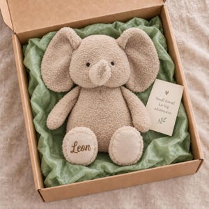 Puede incluir: Un peluche de elefante beige con un acabado de tela rizada texturizada, con el nombre Leon bordado en un pie, colocado en una caja de regalo de cartón forrada con papel de seda verde salvia, junto a una pequeña tarjeta con el texto Small friend for big adventures.