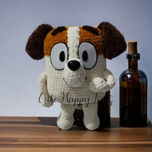 May include: A crocheted stuffed dog toy with brown ears, white fur, and brown and orange patches. The toy has large black eyes and a black nose. The toy is standing on a wooden surface.
