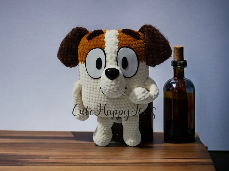 May include: A crocheted stuffed dog toy with brown ears, white fur, and brown and orange patches. The toy has large black eyes and a black nose. The toy is standing on a wooden surface.