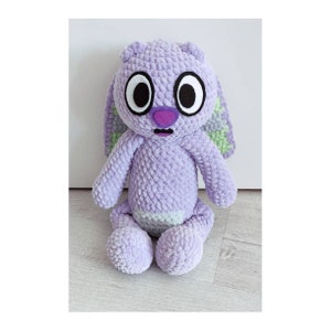 May include: A plush toy made of lavender-colored yarn with a textured, looped finish. It features large, round eyes with black pupils and white sclera, a purple nose, and a small open mouth. The ears have light green accents on the inner side.