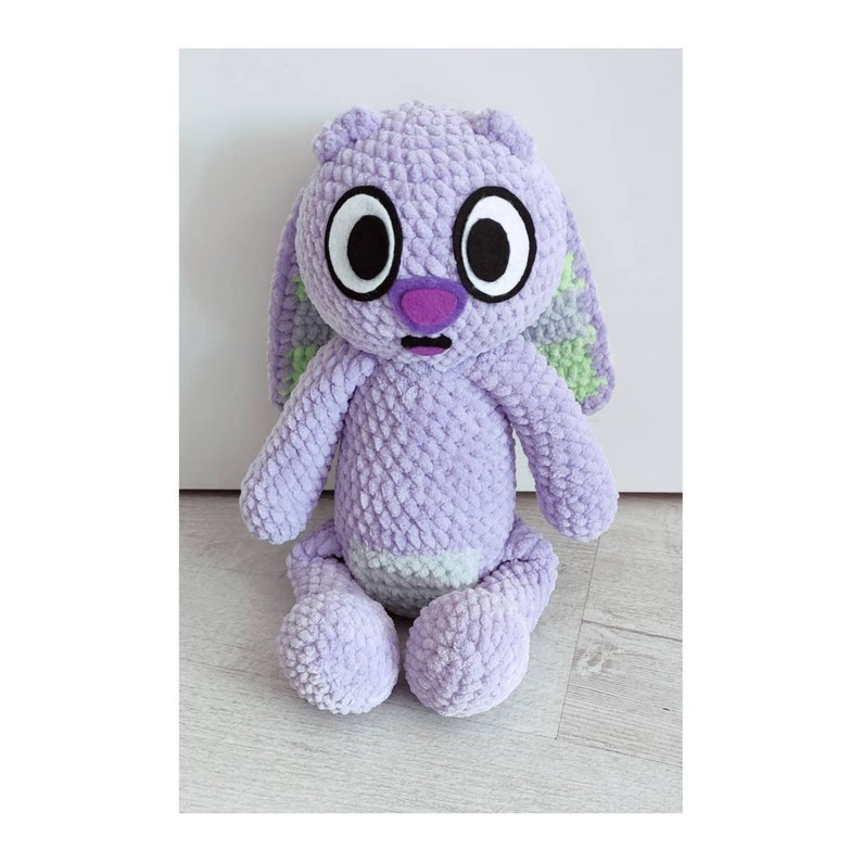 May include: A plush toy made of lavender-colored yarn with a textured, looped finish. It features large, round eyes with black pupils and white sclera, a purple nose, and a small open mouth. The ears have light green accents on the inner side.