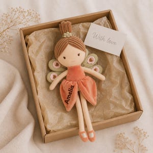 Op de afbeelding: Een pluche poppenfee met bruin corduroy haar, een perzikkleurige fluwelen jurk en saliegroene vleugels met roze hartjes. De naam Amelia is op de jurk geborduurd. Een wit kaartje met de tekst With love zit in de geschenkdoos.