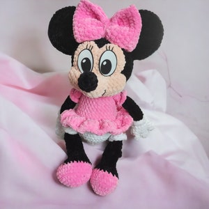 May include: A handmade plush toy of a cartoon mouse character. The toy has a black head with large ears, a pink bow, and a pink dress. The mouse has black legs and feet, and white gloves. The background is a soft pink.