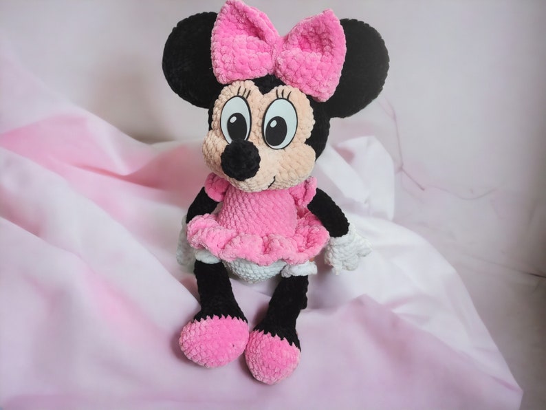May include: A handmade plush toy of a cartoon mouse character. The toy has a black head with large ears, a pink bow, and a pink dress. The mouse has black legs and feet, and white gloves. The background is a soft pink.
