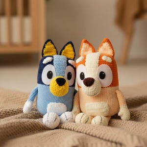 Puede incluir: Dos peluches de perro hechos a mano con ganchillo. Uno es azul con nariz y orejas amarillas, el otro es naranja y blanco. Ambos tienen grandes ojos negros y blancos y están sentados sobre una manta beige.