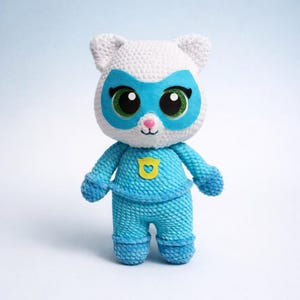 May include: A handmade crochet superhero cat doll. The doll is white with a blue mask, eyes, and outfit. The doll has a yellow shield on the chest. The doll is approximately 8 inches tall.