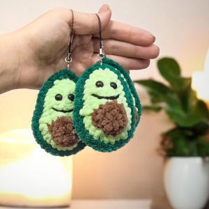May include: Two handmade avocado-shaped earrings. Each earring features a light green exterior, a dark green border, and a brown pit. The avocados have smiling faces with black eyes and a curved mouth. The earrings are held by a black string.