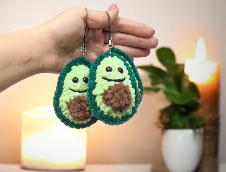 May include: Two handmade avocado-shaped earrings. Each earring features a light green exterior, a dark green border, and a brown pit. The avocados have smiling faces with black eyes and a curved mouth. The earrings are held by a black string.
