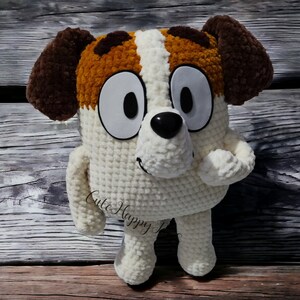 May include: A crocheted plush toy dog with brown ears, white fur, and brown spots. The dog has large white eyes, a black nose, and a happy expression. The toy is sitting on a wooden surface.