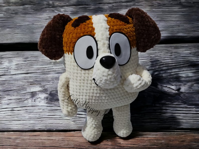 May include: A crocheted plush toy dog with brown ears, white fur, and brown spots. The dog has large white eyes, a black nose, and a happy expression. The toy is sitting on a wooden surface.