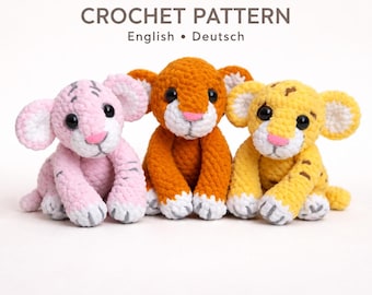 May include: Three crocheted plush animal toys featuring a pink tiger, an orange lion, and a yellow tiger. Each toy has large black eyes, a pink nose, and white paws. The text CROCHET PATTERN English • Deutsch is displayed above the three plush toys.