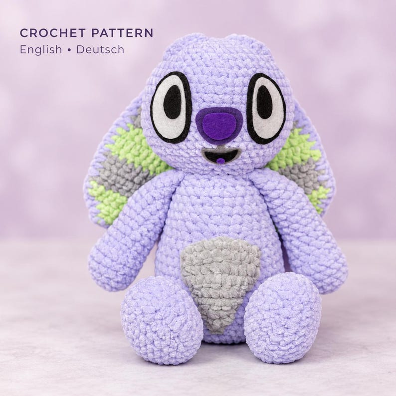May include: A crochet pattern document titled CROCHET PATTERN English &bull; Deutsch featuring a lavender, light green, and gray plush toy with large white and black eyes, a dark purple nose, and a wide mouth, crafted with a textured yarn finish.
