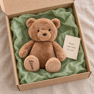 Könnte beinhalten: Ein brauner Plüschteddybär mit gestickten Details an den Füßen, der den Namen Liam auf dem linken Fuß und den Buchstaben J auf dem rechten Fuß trägt. Der Bär liegt in einer Pappschachtel, die mit grünem Seidenpapier und einer Karte ausgelegt ist.