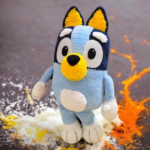 May include: A blue and yellow crocheted stuffed animal dog with white accents. The dog is standing on a brown surface with white and orange powder around it.