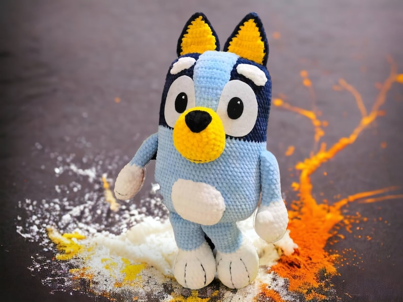 May include: A blue and yellow crocheted stuffed animal dog with white accents. The dog is standing on a brown surface with white and orange powder around it.