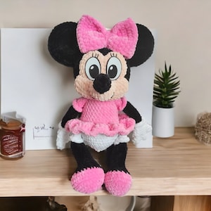 May include: A handmade Minnie Mouse plush doll. The doll features a black head and ears, a pink bow, and a pink dress with a white diaper. The doll is sitting on a wooden surface. The doll is a cute and cuddly toy.