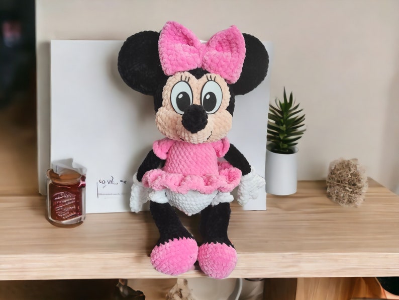 May include: A handmade Minnie Mouse plush doll. The doll features a black head and ears, a pink bow, and a pink dress with a white diaper. The doll is sitting on a wooden surface. The doll is a cute and cuddly toy.