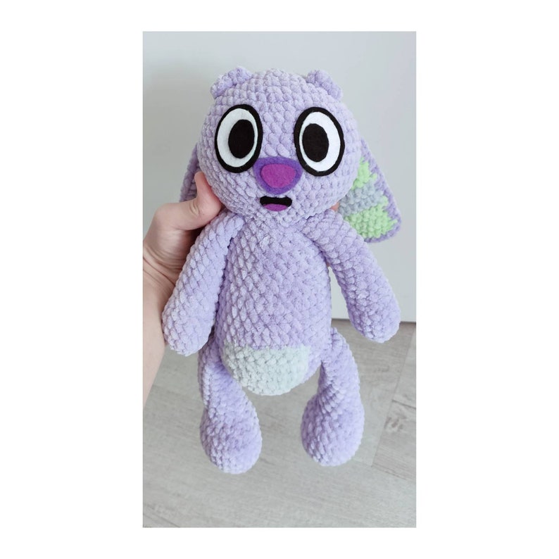 May include: A lavender plush toy with a textured, velvety surface. It features large, round white eyes with black pupils, a purple nose, and a small open mouth. The belly is a lighter shade of lavender, and one ear shows a light green patterned detail.