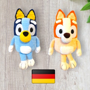 May include: Two crocheted stuffed dog toys. One is blue with white accents and yellow ears. The other is orange with white accents and yellow ears. The toys are sitting on a wooden surface with a German flag in the foreground.