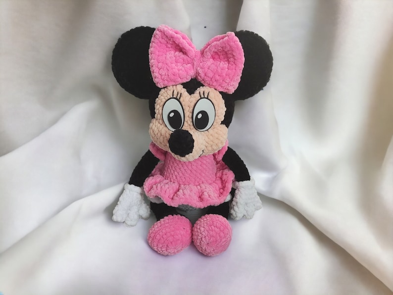 May include: A handmade plush toy of a cartoon mouse character. The mouse has black ears, a pink bow, a pink dress, and white gloves. The toy is made of soft, textured yarn and has large, expressive eyes.