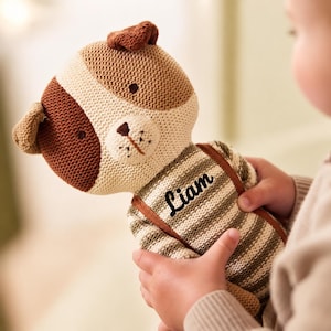 Puede incluir: Un peluche de perro tejido con cara color crema y marrón, ojos y nariz bordados. El cuerpo tiene un patrón de rayas con tirantes marrones y el nombre Liam bordado en letra cursiva negra en la parte superior del pecho.