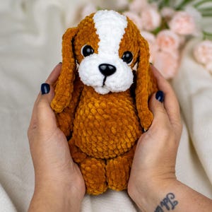 May include: A handmade, plush dog toy in shades of brown, white, and black. The dog has long, floppy ears, a white patch on its head, and a black nose. The toy is soft and cuddly, perfect for children or as a decorative item.