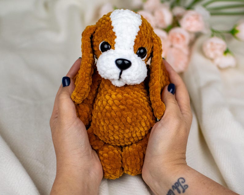 May include: A handmade, plush dog toy in shades of brown, white, and black. The dog has long, floppy ears, a white patch on its head, and a black nose. The toy is soft and cuddly, perfect for children or as a decorative item.