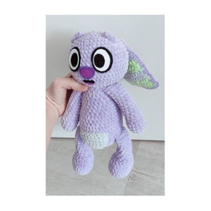 May include: A lavender crochet plush toy with a textured yarn finish. The creature features large white eyes with black pupils, a purple nose, a small open mouth, and long ears with light green inner sections. It has a light green belly patch.