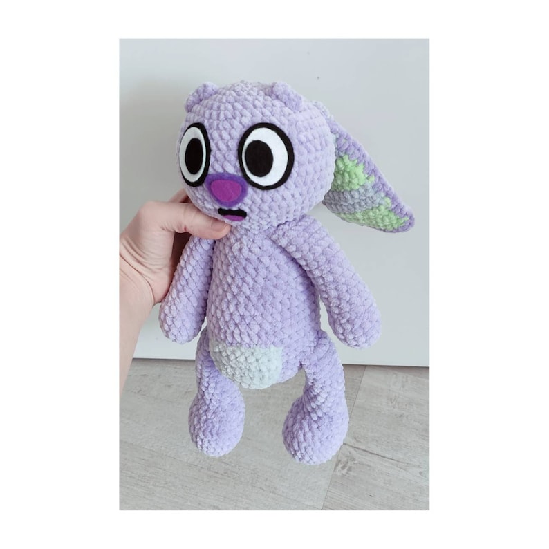 May include: A lavender crochet plush toy with a textured yarn finish. The creature features large white eyes with black pupils, a purple nose, a small open mouth, and long ears with light green inner sections. It has a light green belly patch.