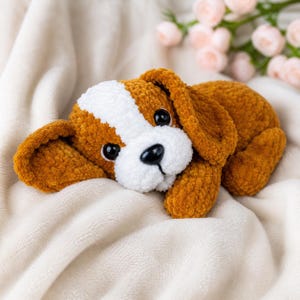 Peut inclure: Un jouet en forme de chien, fait à la main, en peluche marron et blanc, avec des yeux noirs et un nez noir. Le jouet doux est posé sur une couverture crème. Les oreilles du chien sont tombantes et son visage a une expression douce.