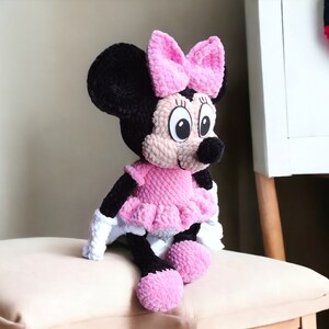 May include: A handmade plush Minnie Mouse doll. The doll features a black head and ears, a pink bow, a pink dress, and white gloves. The doll is sitting on a beige cushion.