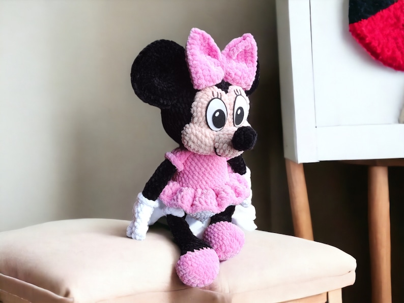 May include: A handmade plush Minnie Mouse doll. The doll features a black head and ears, a pink bow, a pink dress, and white gloves. The doll is sitting on a beige cushion.