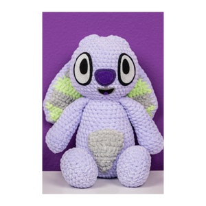 May include: A crochet plush toy featuring a light purple body with a textured yarn finish. The character has large white and black eyes, a dark purple nose, and long ears with light green and grey accents. A grey patch is centered on the belly.