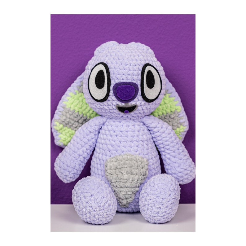 May include: A crochet plush toy featuring a light purple body with a textured yarn finish. The character has large white and black eyes, a dark purple nose, and long ears with light green and grey accents. A grey patch is centered on the belly.