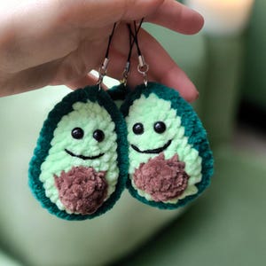 May include: Two handmade avocado-shaped keychains. Each keychain features a light green center, a dark green border, and a brown pit. The avocados have black eyes and smiling mouths. They are attached to black cords.