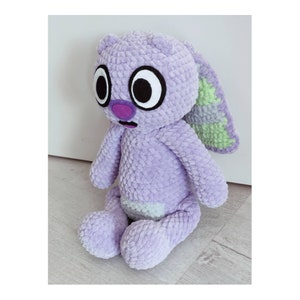 May include: A plush toy made of lavender textured yarn with large white and black eyes, a purple nose, and a small open mouth. The creature has long ears with light green accents on the inner side and a light green patch on its belly.
