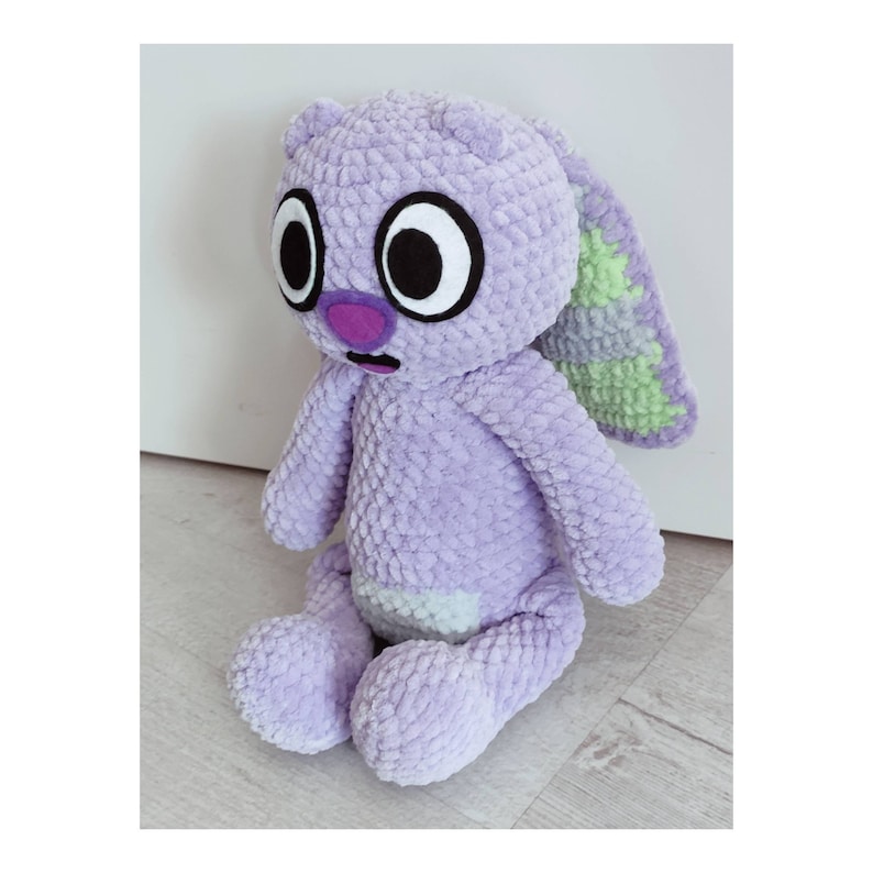May include: A plush toy made of lavender textured yarn with large white and black eyes, a purple nose, and a small open mouth. The creature has long ears with light green accents on the inner side and a light green patch on its belly.