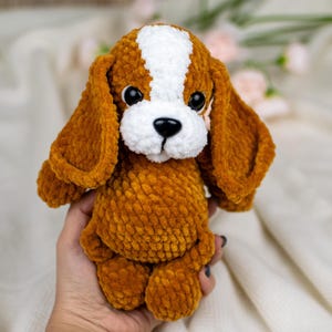 May include: A handmade plush dog toy with brown and white fur. The dog has large, floppy ears, a white muzzle, and black eyes. The toy is made of a soft, textured material and is designed to resemble a beagle puppy.