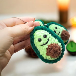 May include: Handmade crochet avocado keychains. Each keychain features a light green body with a dark green border, a brown seed, and a smiling face. The keychains are small and lightweight.